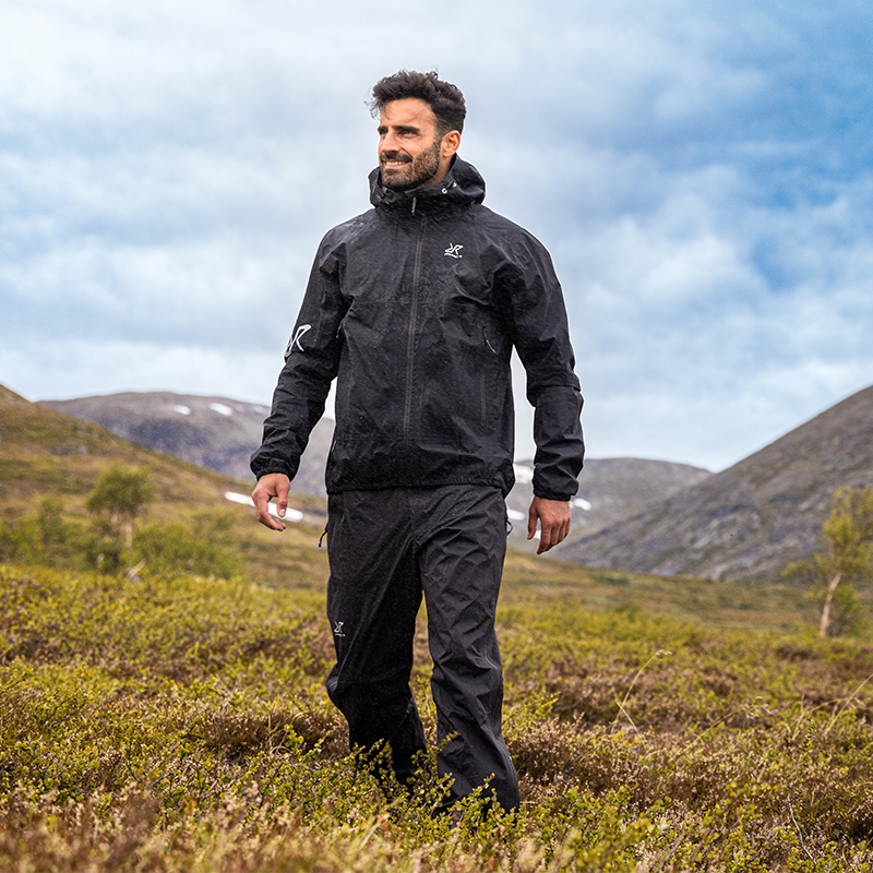 Vandrer i sort skaljakke og regnbukser går gennem lyng mellem fjelde